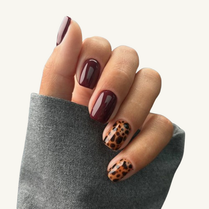A hand wearing a set of short square press-on nails with a mix of solid burgundy and brown leopard print patterns.