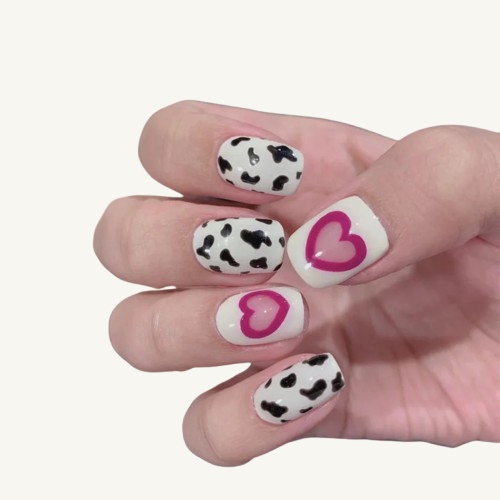 A hand wearing Oval press-on nails with a mixed design of black and white cow print and pink jelly-effect heart patterns.