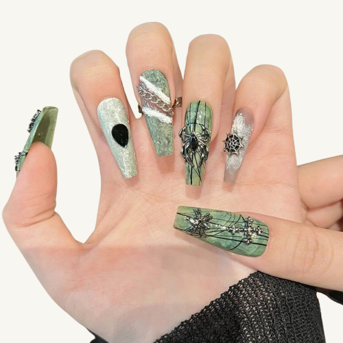 Hands wearing sage green punk-style press-on nails, with 3D metallic spiders, chains, and hand-painted lines.