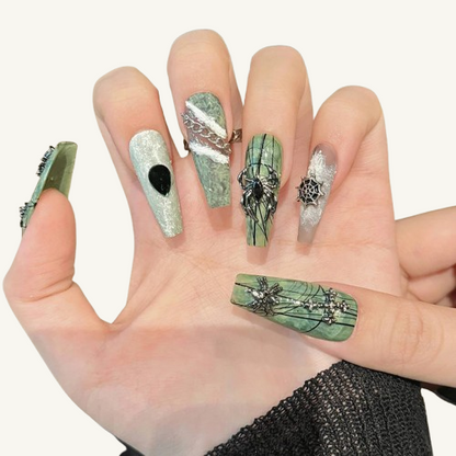 Hands wearing sage green punk-style press-on nails, with 3D metallic spiders, chains, and hand-painted lines.