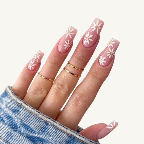 A hand wearing Coffin press-on nails with white daisy patterns on a nude-pink base, paired with gold rings.