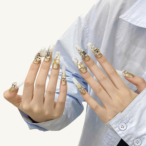 A set of long square press-on nails with a gold glitter French design, featuring luxury gemstone inlays on a grey-blue base.