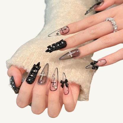 A set of long stiletto gothic press-on nails in a black and white theme, adorned with 3D bows, crosses, chains, and rhinestones.