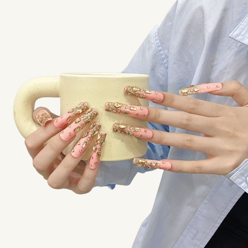 A set of long square press-on nails with a nude pink base, adorned with gold glitter and 3D gold teardrop gems at the tips.