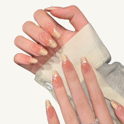 A set of handmade oval press-on nails, peach ombre, with liquid-textured gold French tips and adorned with iridescent flakes.
