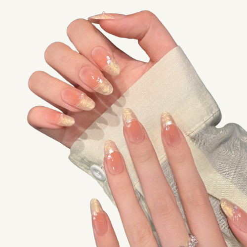 A set of handmade oval press-on nails, peach ombre, with liquid-textured gold French tips and adorned with iridescent flakes.
