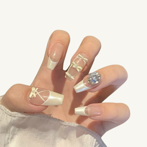 A hand wearing long square press-on nails with a pearl white French design, cream bows, and a large heart-shaped diamond accent.