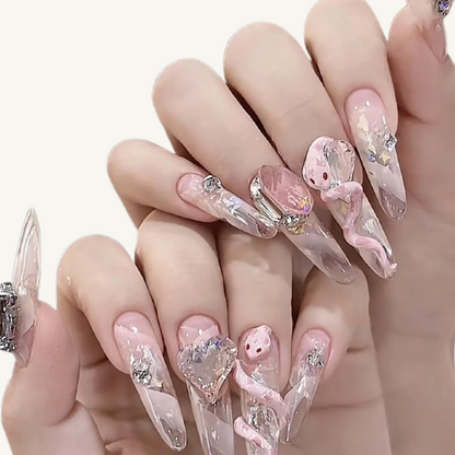 Hands wearing long coffin nails with a nude pink base, featuring handmade 3D pink snakes, large pink crystals, and heart-shaped gems.