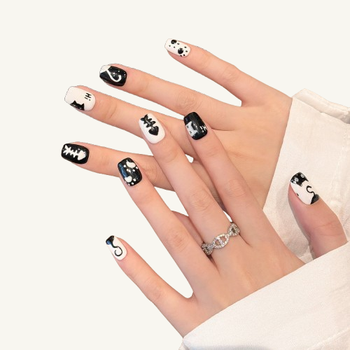 A hand wearing black and white graffiti cat-themed press-on nails featuring cat silhouettes, paw prints, fishbones, and polka dots, while holding a mug.