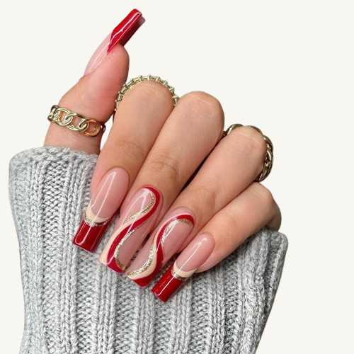 A hand wearing long square press-on nails with a red, cream, and gold glitter swirl design, adorned with gold rings.