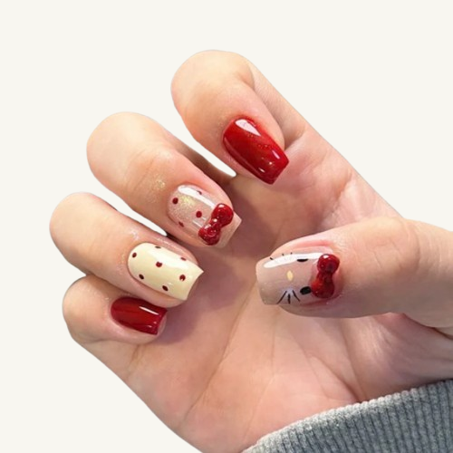 A hand wearing a set of short square press-on nails with designs including a 3D red bow, red polka dots, and a cat whisker pattern.