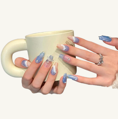 A set of wave and sand textured press-on nails in light blue glitter and blush pink, with a mermaid tail and rhinestone charms.
