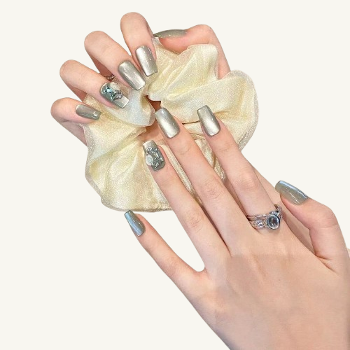 A set of silver-green cat-eye press-on nails in a square shape, with one nail accented by a blue gemstone and a white rose.