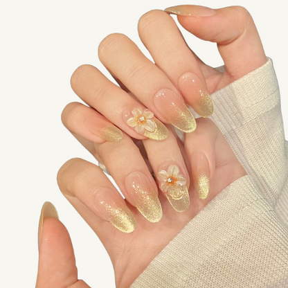 A set of handmade press-on nails, peach nude to gold glitter gradient, and adorned with a 3D glass-textured flower.