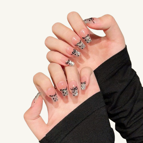 A set of handmade coffin press-on nails with a nude gradient base and silver glitter tips featuring black polka dots and hand-painted bows.
